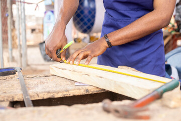 A carpenter wears a blue apron while working with tools, measuring and marking a wooden plank. Workshop environment in the background with visible tools and raw building materials.