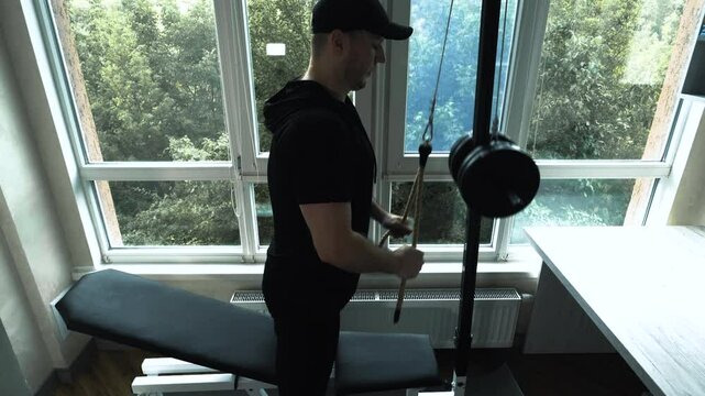 Pumping triceps arms on a sports simulator in the apartment. A young man trains his triceps by doing rope rows. Workout at home. Slow motion
