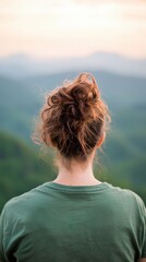 Fototapeta premium A contemplative person with curly hair stands on a mountain, gazing at a serene landscape with soft hues of sunrise or sunset in the background.
