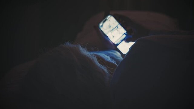 woman suffers from insomnia lying in bed using smart phone before sleep, dark night room. light from affects eyes face. lady scrolling social media app Phone screen holding mobile in hands. old age
