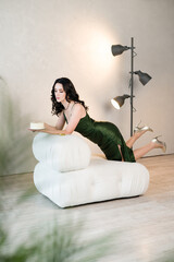 A stylish woman in a dark green dress leans on a contemporary white sofa, holding a small cake. The...