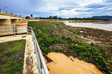 Obraz premium View of irrigation canal with weeds and debris flowing together with strong flowing dirty brown water due to soil and mud from forest after heavy rains and floods. La Nina crisis, Water pollution.