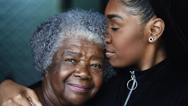 Tender loving moment of African descent granddaughter kissing elderly South American grandmother in the forehead in caring embrace with arm around shoulder, close-up faces