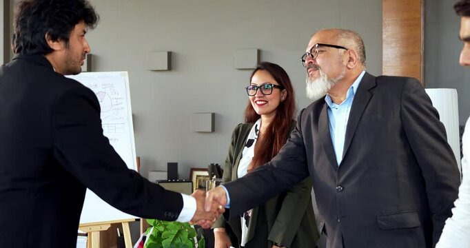 Indian Asian young business professional or businessman confidently shaking hands across a table with a colleague after finalizing a significant business deal or new project in modern corporate office
