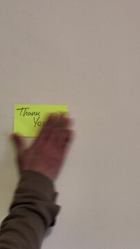 A yellow sticky note with the word "Thank you" handwritten in simple handwriting, stuck on a wall