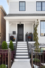 A black front door on a suburban grey and white house with plants on a small front porch and black railing.