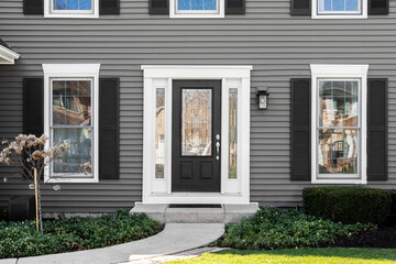 A front door detail on a dark grey suburban home with a black and glass front door, white windows, and a concrete sidewalk.