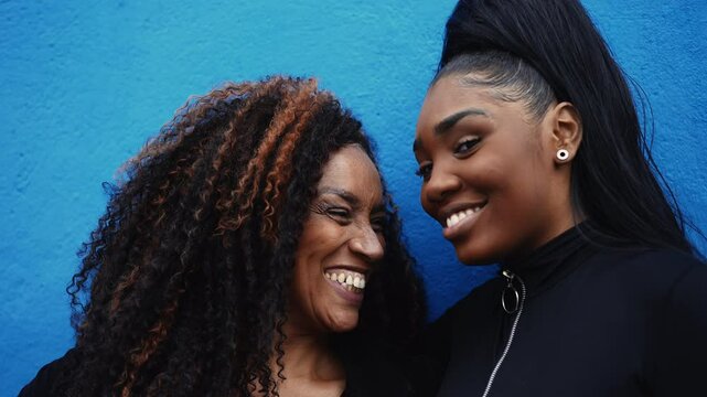 African American adolescent daughter kissing mother in the forehead showing family love and tenderness standing on blue wall. Parent and child bonding affectionate moment