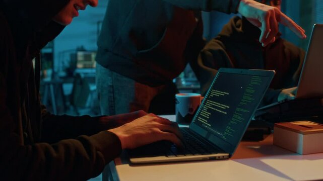 Tilt down view of young male fraudster engaging in illegal activities using laptop while two hackers working together in background