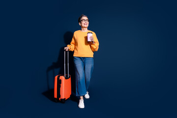 Full body photo of mature woman traveling suitcase drink coffee walk look empty space wear yellow clothes isolated on dark blue background