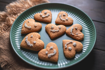 Delicious heart-shaped gingerbread cookies decorated with a smiley face made of white sugar pearls are arranged on a green plate placed on a dark wooden table with a rustic burlap tablecloth