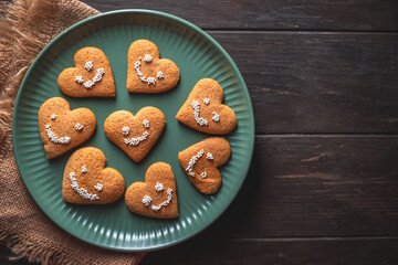 Delicious heart-shaped gingerbread cookies decorated with a smiley face made of white sugar pearls are arranged on a green plate placed on a dark wooden table with a rustic burlap tablecloth