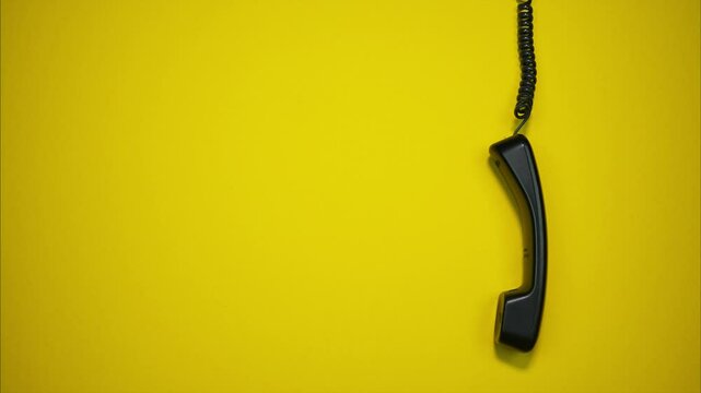 An old black telephone receiver hanging on a cord on yellow background. Stop motion