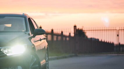 Sleek vehicle parked roadside, gleaming headlights illuminating dramatic sunset sky with vibrant orange pink gradient