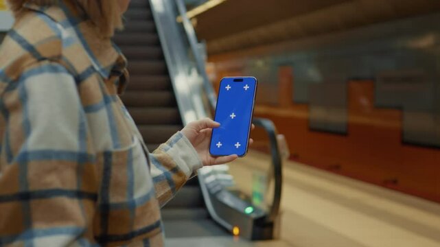 Woman showing a smartphone with chroma key blue screen and tracking markers while standing still on an escalator in a subway station