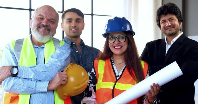 Indian Asian group of civil engineers and architects wearing fluorescent safety jackets, hard hats, holding rolled blueprints, standing confidently with folded arms, smiling, looking at camera
