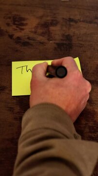 A person writes a thank you note on a piece of paper. The note is on a wooden table and the person uses a marker to write it. The scene is positive and thankful.