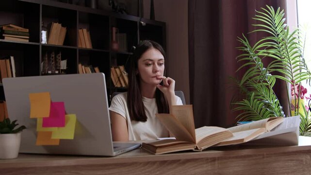 Thinking girl is preparing for her bachelor's thesis defense. Student at an office table with books, notebooks, computer is writing her master's thesis.Young woman is studying in a prestigious library