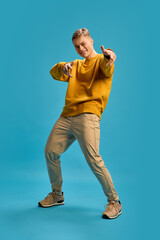 Naklejka premium Man pointing forward with playful smile, leaning slightly, engaging expressively with energetic gestures against blue studio background. Concept of human emotions, beauty, self-expression.