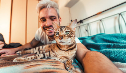 Owner playing with cat relaxing on the bed at home - Happy man and bengal cat taking selfie picture indoor - Domestic animal and people life style concept