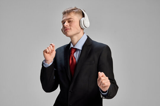 Overjoyed, relaxed young man, dressed formal attire, dancing while listening music in headphones against grey studio background. Concept of human emotions, business, self-expression. - Powered by Adobe