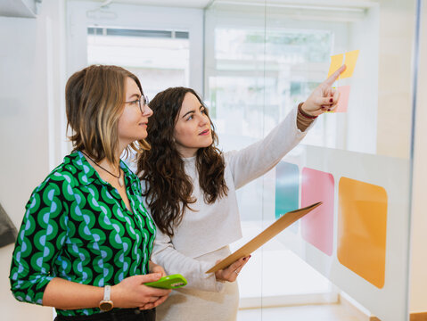 Two professional women brainstorming in a modern coworking space. One points at sticky notes on a glass wall while holding a clipboard, reflecting teamwork and strategy.