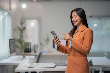 Young Asian businesswoman wearing suit and badge, holding smartphone, pointing finger at screen, smiling, standing in bright modern office, desk, computer, coffee cup, documents, windows