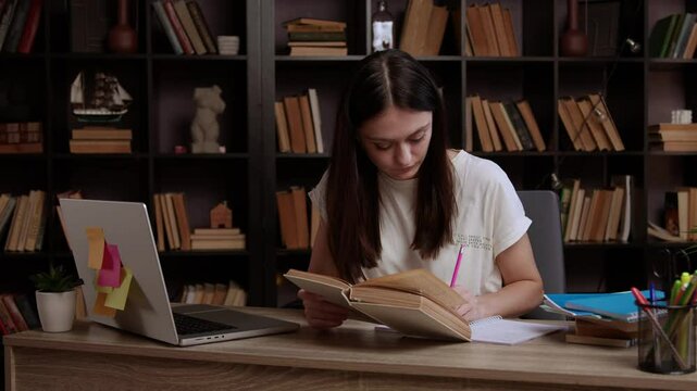 Young student studying in a modern library. Brunette is reviewing information from book and computer and making notes on paper. Girl is intensively preparing for a project for college