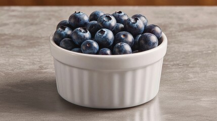 A white bowl filled with fresh, plump blueberries sits on a countertop, showcasing their vibrant color and texture.