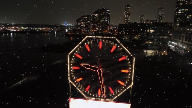 Patriotic flag on lighting Colgate clock on river shore of Hudson River. New Jersey city during snowfall at night. Aerial view. Skyline with lighting skyscraper.