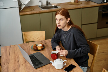 A young guy with long hair sits at a stylish table, sipping tea and enjoying pastries in his home.