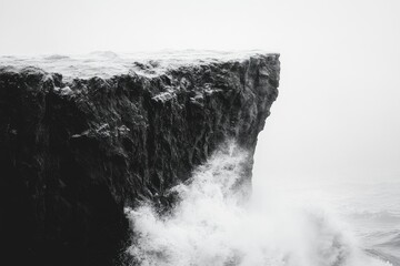 Snow-dusted cliff dramatically battered by powerful ocean waves in a misty, monochrome scene.