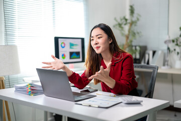 A woman in a red shirt is sitting at a desk with a laptop and a monitor