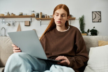 A young man with long red hair is focused on his laptop while relaxing on a couch.