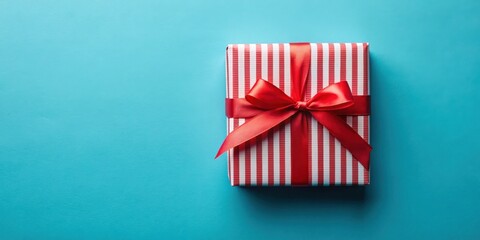 Fototapeta premium A single red and white striped gift box with a bright red ribbon bow sits on a light blue background