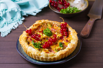 Quiche with chunks of ham, creamy brie and vegetables on blue plate on brown wood background