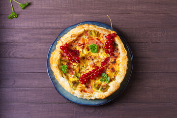 Quiche with chunks of ham, creamy brie and vegetables on blue plate on brown wood background. View from above, top view