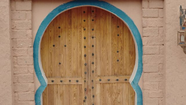 Traditional Moroccan wooden door with ornate metal studs in a peach-colored clay brick wall with a blue arch. A decorative metal lantern hangs beside the door. travel, culture,