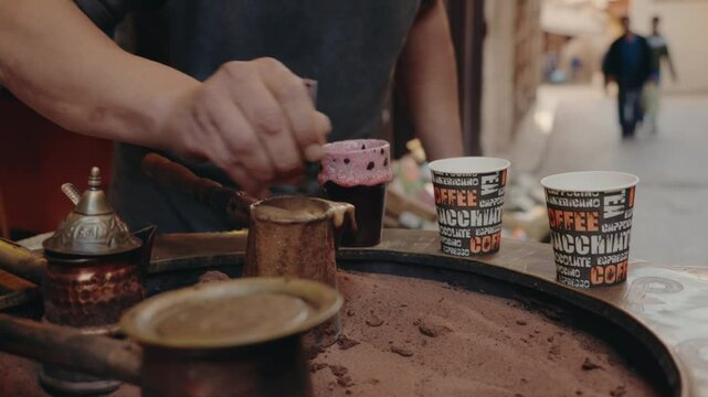 A man prepares traditional Moroccan coffee in a bustling market. He stirs the coffee in a brass pot nestled in hot sand, surrounded by ornate copper pots.The rich aroma fills the air, Moroccan culture