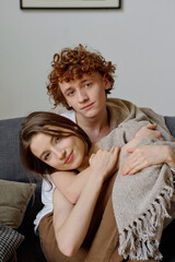 Vertical portrait of young adult couple sitting on sofa in living room, woman smiling lightly while...