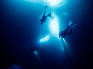 silhouette of unrecognizable scuba divers with flashlights swimming in narrow dark cave with rough rocky formations undersea in deep clear water, concept of active leisure and underwater adventure