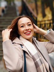 A beautiful young Caucasian woman in her 40s wearing a coat and headscarf in the fall in a park sits on the stairs. Autumn woman portrait. Face with a smile
