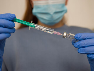 Doctor wearing blue gloves and surgical mask carefully filling syringe with vaccine from glass...