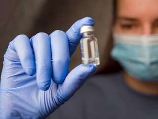 Doctor wearing surgical mask and blue latex gloves holding medical vial containing clear liquid, carefully preparing for medical procedure, emphasizing safety with medical vial in focus. 