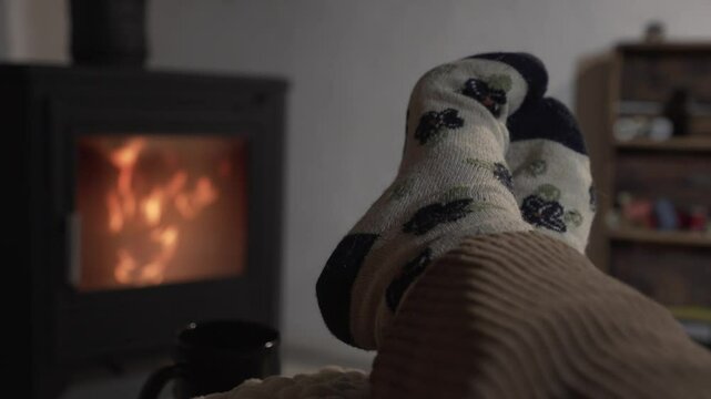 horizontal video of bare feet of a woman with patterned sock resting on a pouf in front of a wood fireplace