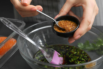 Woman adding mustard into bowl with soy marinade at dark gray table, closeup