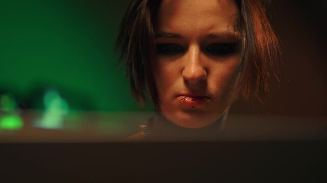 Exhausted female hacker or programmer working with computer in night, portrait. Cybercrime and cybersecurity, woman with tattoos and piercing, alternative style of appearance, possessed black eyes