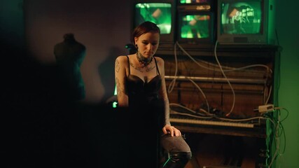 Depressed offbeat young woman sitting alone in strange studio or basement. Static portrait of frustrated freaky lady with tattooed body and piercing on face, abandoned and misunderstood by society