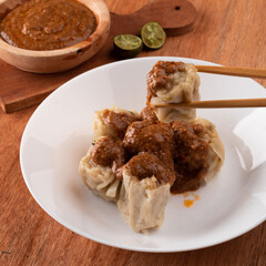 Steamed dumplings topped with a rich, spicy sauce are presented on a white plate, accompanied by lime slices on the side