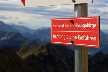 Red warning sign in the Alps with German inscription: Here you are in the high mountains, Attention alpine dangers, hiking in high mountains is dangerous for inexperienced people